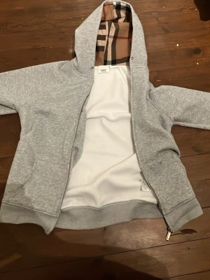 Grå hoodie från Burberry med dragkedja helt ny aldrig använd för den va för liten hör av dig vid frågor - Snygg grå hoodie från Burberry med klassiskt rutigt foder i huvan och dragkedja framtill. Tillverkad i mjukt bomullsmaterial och har ribbade muddar. Perfekt för en chill och stilren look. Ikonisk Burberry-detalj på dragkedjan.