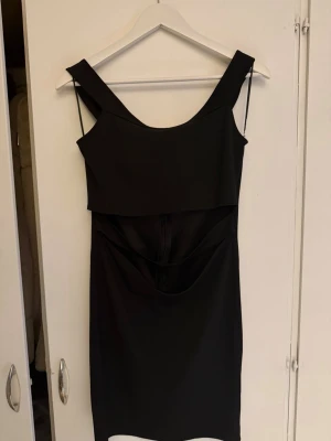 Svart kort klänning med cut out-detalj - Snygg svart kort klänning med breda axelband och trendig cut out-detalj i midjan. Klänningen har en figurnära passform och är perfekt för dig som vill ha en stilren men ändå edgy look. Tillverkad i ett mjukt och stretchigt material.