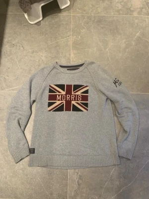 Lite schysstare Grå stickad tröja från Morris - Snygg grå stickad tröja från Morris med Union Jack-flagga och logga framtill. Tröjan har rund hals, ribbade muddar och diskret broderad text på ärmen. Perfekt för dig som gillar klassisk men trendig stil.