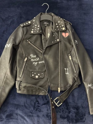 Svart skinnjacka med nitar och tryck - Svart skinnjacka med coola nitar på axlar och krage, bälte i midjan och flera vita tryck. Storlek S. Jackan har dragkedjor, fickor och en edgy bikerstil.
