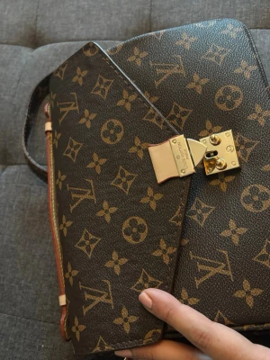 Monogram axelväska (knappen är trasig)  - Snygg axelväska i klassiskt brunt monogrammönster med guldiga metalldetaljer och lås. Väskan har justerbar axelrem, bakficka med dragkedja och stilren design. Perfekt för dig som gillar lyxiga accessoarer och tidlös stil. Knappen för att stänga väskan är trasig och öppnas hela tiden 