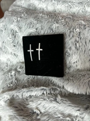 Silvriga korsörhängen med stenar från Edblad - Säljer ett par silvriga örhängen i form av kors, dekorerade med små gnistrande stenar. Perfekta för dig som gillar stilrena och trendiga accessoarer med lite edge. Snygga att matcha med flera andra smycken.