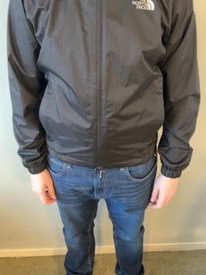 Svart vindjacka från The North Face - Svart vindjacka från The North Face med dragkedja framtill och justerbar huva. Jackan har en minimalistisk design med logga på bröstet och elastiska muddar vid ärmslut och midja. Perfekt för blåsiga dagar och enkel att matcha med jeans.