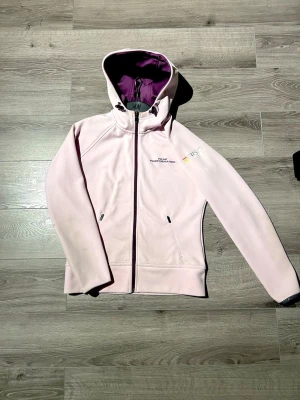Ljusrosa vindjacka Peak Performance - Ljusrosa vindjacka från Peak Performance med dragkedja framtill och huva. Jackan har broderad logga på bröstet och R&D-detalj på ärmen. Insidan är fodrad i lila och jackan har två fickor med dragkedja.