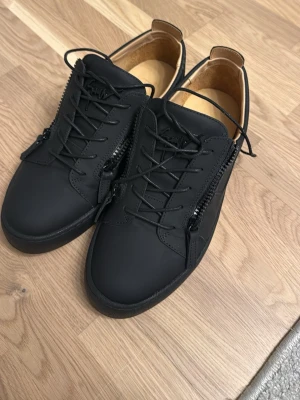 Svarta sneakers från Giuseppe Zanotti - Snygga svarta sneakers i läder från Giuseppe Zanotti med mattsvart finish, svarta snören och dragkedjor på sidorna. Skorna har låg profil och diskret logga bak på sulan. Perfekta för en stilren och modern look.