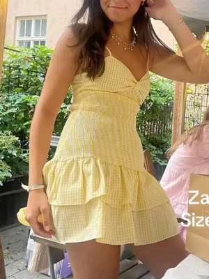 Gul rutig klänning - Superfin gul och vit rutig kortklänning med volanger och smockad midja. Klänningen har smala axelband, omlottdetalj vid bysten och knyt i ryggen. Perfekt för somriga dagar och ger en fräsch look.