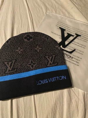 svart och blå mössa - Snygg svart mössa med klassiskt monogrammönster i grått och en blå rand. Märkets logga är broderad i blått framtill. Perfekt accessoar för att lyfta din streetwear-look.