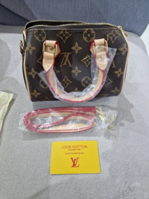 Louis Vuitton Monogram handväska - Säljer en klassisk Louis Vuitton handväska i brunt monogrammönster med beige och röda detaljer. Väskan har guldfärgade metalldetaljer, avtagbar axelrem och dragkedja. Ikonisk LV-logga och blommönster på slitstarkt canvasmaterial.