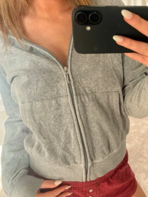 Grå Juicy Couture hoodie med dragkedja - Säljer en grå hoodie från Juicy Couture med dragkedja framtill och huva. Baksidan har glittrande Juicy-text i stenar. Hoodien har bred mudd i midjan och vid ärmsluten samt två fickor fram. Perfekt för en chill och trendig look.