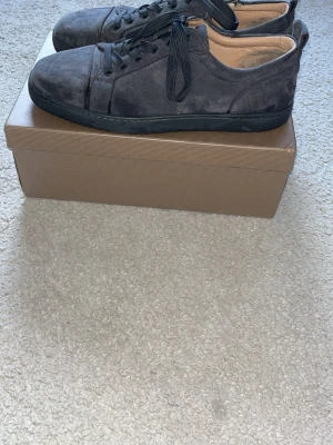 Gråa sneakers med röd sula - Snygga gråa sneakers i mocka från Christian Louboutin med klassisk röd sula och svarta skosnören. Lågt skaft och diskret design som passar till många olika outfits. Finns en defekt men som inte syns då man väl har på sig dem och det påverkar inte skorna alls.  R€P e dem också