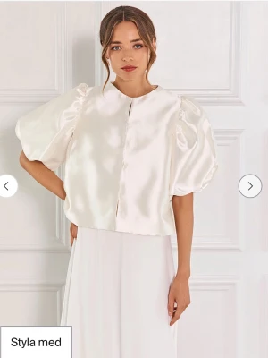 Malina -Vit satinblus med puffärm - Stilren vit blus i glansigt satinmaterial från By Malina  med korta puffärmar och rund halsringning. Blusen har en rak, något boxig passform och dold knäppning framtill. Färgen är ivory. Nypris 2200 kr. 