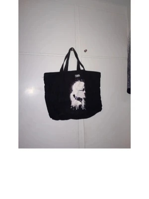 Svart shoppingväska Karl Lagerfeld - Cool svart shoppingväska från Karl Lagerfeld med ett stort vitt tryck av Karl Lagerfelds ikoniska profil på framsidan. Väskan har dubbla handtag och är rymlig, perfekt för att bära med sig allt man behöver. Materialet är tyg och designen är riktigt trendig.