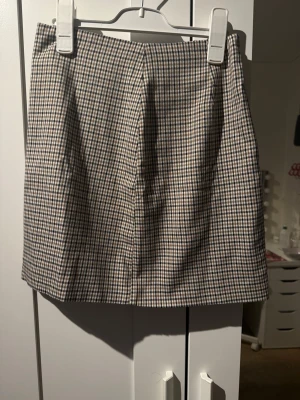 Rutig kortkjol i beige och svart - Trendig kortkjol med rutigt mönster i beige, svart och vitt. Kjolen har en rak passform och klassisk midja, perfekt att styla med en stickad tröja eller t-shirt. Materialet känns mjukt och bekvämt, och kjolen har en tidlös look.