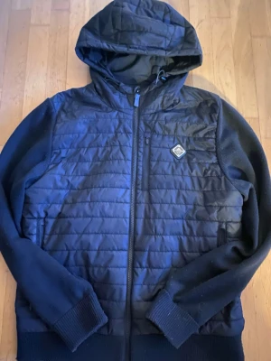 Svart cardigan från J.Lindeberg - Snygg svart vindjacka från J.Lindeberg med quiltad front, huva och ribbade muddar. Jackan har dragkedja framtill och en diskret logotyp på bröstet. Passar M och L. 