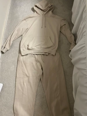 Beige mjukis-set  - Snyggt och bekvämt loungewear-set i beige med matchande hoodie och mjukisbyxor. Hoodien har känguruficka och dragsko i huvan, byxorna har resår och dragsko i midjan. Perfekt för chill dagar hemma.