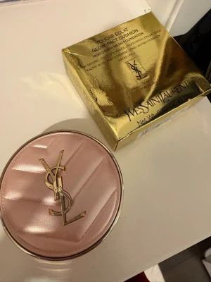 YSL Touche Éclat Glow-Pact Cushion - Säljer en YSL Touche Éclat Glow-Pact Cushion foundation i färgen 10 Porcelain. Kommer i en lyxig rund rosa dosa med guldfärgade detaljer och YSL-logga. Ger naturlig lyster, hög täckning och långvarig återfuktning. SPF 23. Förpackningen är guldig och elegant.