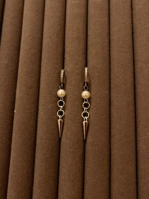 Silvriga örhängen med pärlor och spikes - Säljer ett par trendiga örhängen i silverfärgad metall med vita pärlor och spetsiga spikes. Örhängena har små ringar som länkar ihop pärlan och spiken, vilket ger en cool och edgy look. Perfekta för dig som vill sticka ut lite extra.
