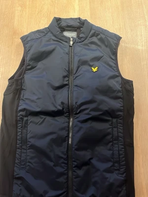 Svart och blå  dunväst från Lyle & Scott. (Storlek S) - Snygg svart och blå  dunväst från Lyle & Scott med gul logga på bröstet. Västen har dragkedja framtill, två fickor och är tillverkad i ett glansigt material. Perfekt för lager-på-lager och streetwear-stil.