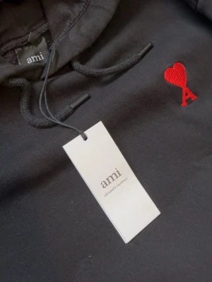 Svart hoodie från Ami med rött hjärta - Snygg svart hoodie från Ami Alexandre Mattiussi med klassisk huva och dragsko. På bröstet finns Ami:s ikoniska röda hjärta med bokstaven A broderat. Hoodien har känguruficka och ribbade muddar. Perfekt för en chill och trendig look.