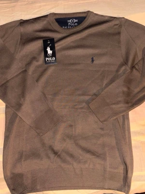 Brun stickad tröja Polo Ralph Lauren - Snygg brun stickad tröja från Polo Ralph Lauren med rund halsringning och diskret broderad logga på bröstet. Tröjan har långa ärmar och ribbade muddar vid ärmslut och nederkant. Perfekt för lager på lager och stilren look.