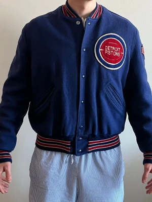 Vintage 90s Detroit Pistons DeLONG Varsity Jacket USA Made NBA Bad Boys Era - Äkta vintage Detroit Pistons varsity-jacka från DeLONG. Tillverkad i USA i kraftig ullmodell från sent 80-tal / tidigt 90-tal (Bad Boys-eran). Broderade patches framtill, på ärmen samt stor ryggpatch. Mycket fint vintageskick.                                     Modell på bild: 183cm, 81kg