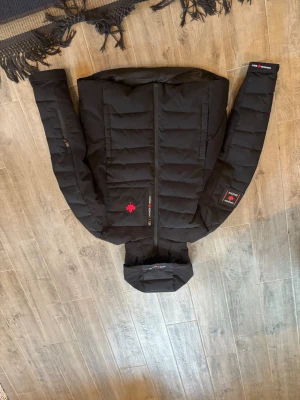 Svart pufferjacka från Save The Duck - Snygg svart pufferjacka från Save The Duck med huva och dragkedja framtill. Jackan har diskreta logopatchar på ärmen och bröstet samt två sidofickor med dragkedja. Tillverkad i ett slätt, quiltat material som ger en modern look. Den är inprinsip som ny