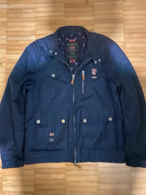 Mörkblå Morris vindjacka - Mörkblå vindjacka från Morris med emblem på bröstet, dragkedja med läderdetaljer, två stora fickor med knappar och en bröstficka med dragkedja. Jackan har mönstrat innerfoder och klassisk krage. Materialet är lätt och passar perfekt för blåsiga dagar.