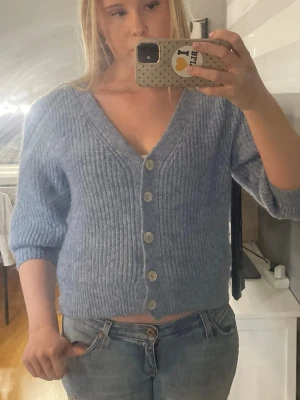 Blå stickad kofta med knappar - Mysig blå stickad kofta med v-ringning och stora ljusa knappar framtill. Koftan har ribbstickat mönster och trekvartsärmar, perfekt att slänga på sig över en t-shirt. Materialet ser ut att vara mjukt och fluffigt.