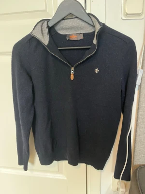 Mörkblå halvzip-tröja från Morris - Mörkblå stickad tröja från Morris med halv dragkedja och liten broderad logga på bröstet. Tröjan har ribbade muddar och krage med grå insida. Perfekt för lager-på-lager och snygg till jeans.