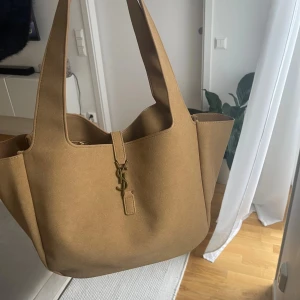 Beige axelväska  - Snygg och rymlig axelväska från i beige mocka med ikoniskt spänne i guld framtill. Väskan har breda handtag och stilren design som gör den enkel att matcha med olika outfits. Perfekt för dig som gillar lyxiga accessoarer med klassisk touch.