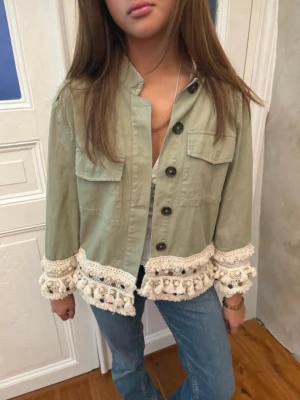 Grön jacka med virkade detaljer - Stilren ljusgrön jacka med virkade och tofsprydda detaljer längs ärmslut och nederkant. Jackan har knappar framtill, två bröstfickor och en rak, croppad passform. Perfekt för dig som vill sticka ut med en unik look.