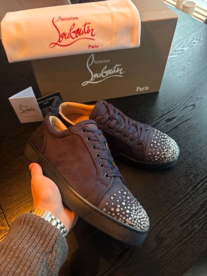 Christian Louboutin sneakers - Snygga mörkblå sneakers från Christian Louboutin i mocka med ikoniska röda sulor och silverfärgade nitar på tån. Skorna har lågt skaft, matchande mörkblå snörning och exklusiv känsla. Perfekta för dig som vill sticka ut med stilrena detaljer.
