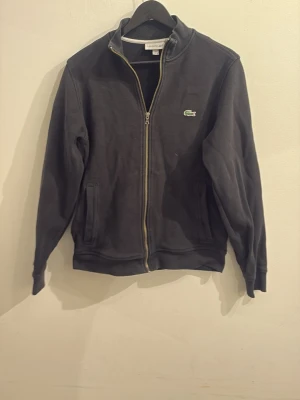 Lacoste zip up - Snygg zip up från Lacoste med dragkedja framtill och klassisk krokodillogga på bröstet. Tröjan har två fickor och ribbade muddar vid ärmslut och nederkant. Perfekt för en avslappnad och stilren look.
