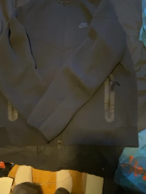 Svart Nike Tech Hoodie - Svart hoodie från Nike med dragkedja framtill och två fickor med dragkedja.  Perfekt för en avslappnad stil 