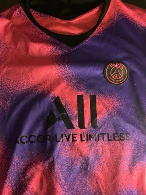 PSG matchtröja rosa/lila Nike - Säljer en snygg PSG matchtröja i rosa och lila med svart logga och texten 'Accor Live Limitless' på bröstet. Tröjan har klubbmärket Paris Saint-Germain på vänster sida och V-ringning. Perfekt för fotbollsfans!