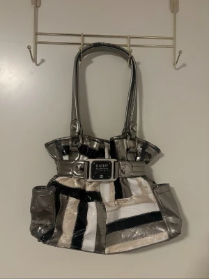 Randig handväska från Kathy Van Zeeland - Snygg handväska från Kathy Van Zeeland med metallicdetaljer och randigt mönster i silver, svart, vitt och beige. Väskan har två axelband, dekorativt spänne framtill och flera fack. Perfekt för dig som gillar statement-accessoarer.