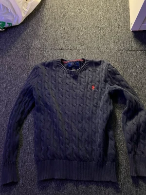 Mörkblå kabelstickad tröja Polo Ralph Lauren - Kabelstickad mörkblå tröja från Polo Ralph Lauren med rund halsringning och ribbade muddar. Tröjan har det klassiska röda Polo-logot på bröstet och är tillverkad i mjukt material. Perfekt för dig som gillar tidlös och stilren design.