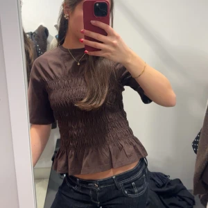 Brun smockad topp  - Brun topp med smockad midja och korta ärmar. Modellen har en lätt volang nedtill och rund halsringning. Verkligen så snygg o classy, aldrig använd, endast testad💗
