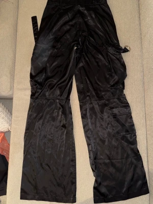 Svarta cargobyxor i satin från Bershka - Snygga svarta cargobyxor i glansigt satinmaterial från Bershka. Byxorna har vida ben, flera fickor på sidorna och coola detaljer som band och metallring. Perfekta för en trendig streetstyle-look.