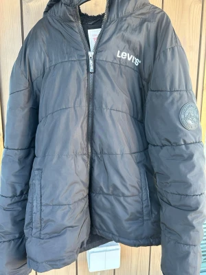 Svart pufferjacka från Levi's - Svart pufferjacka från Levi's med huva och dragkedja framtill. Jackan har två fickor, broderad logga på bröstet och ett runt märke på ärmen. Tillverkad i ett glansigt, vadderat material som håller värmen under kalla dagar.