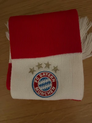 FC Bayern München halsduk - Stickad halsduk i rött och vitt med breda ränder och broderad FC Bayern München-logga samt fyra guldstjärnor. Halsduken har vita fransar i ändarna och är perfekt för dig som vill visa ditt stöd för laget. Materialet är mjukt och värmande.