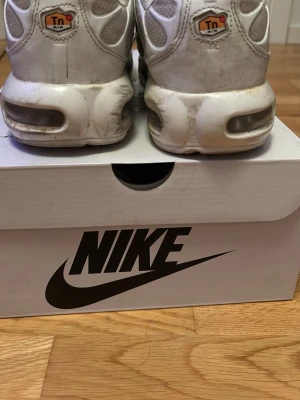 Nike Air Max Plus vita sneakers - Nike Air Max Plus sneakers i vitt med silvriga detaljer och Tn-logga på hälen. Skorna har en sportig design med synlig Air Max-dämpning i sulan och mesh- och syntetmaterial på ovandelen. Perfekta för dig som gillar klassiska och stilrena sneakers.
