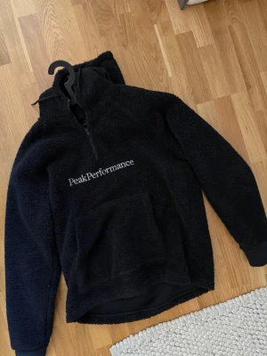 Svart fleece från Peak Performance - Mysig svart fleece med halv dragkedja och stor magficka från Peak Performance. Tröjan har broderad logga på bröstet och är tillverkad i fluffigt material som håller dig varm. Perfekt för lager-på-lager och chill dagar.