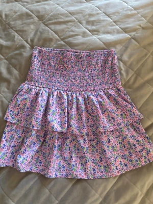 Blommig volangkjol från Vero Moda - Supersöt kort kjol från Vero Moda med blommigt mönster i rosa, blått, grönt och lila. Kjolen har bred smock i midjan och dubbla volanger för extra flowy känsla. Perfekt för vår och sommar, matcha med en enkel topp för en fräsch look. Storlek S 