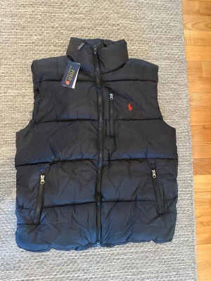Svart dunväst från Polo Ralph Lauren - Stilren svart dunväst från Polo Ralph Lauren med hög krage, tre dragkedjefickor och klassisk röd logga på bröstet. Västen är quiltad och har en sportig look, perfekt att slänga över en hoodie eller tröja. STORLEK L 