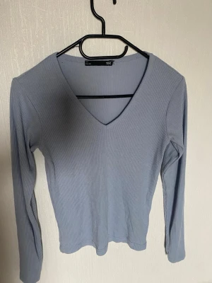 Ljusblå ribbad långärmad topp - Säljer en ljusblå ribbad långärmad topp med v-ringning. Toppen har en smal passform och är tillverkad i ett mjukt, stretchigt material som sitter skönt på kroppen. Perfekt att matcha med jeans eller kjol för en enkel och snygg look.