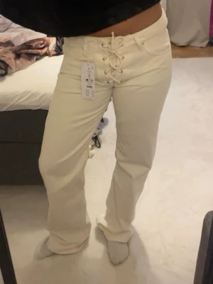 Vita jeans med snörning i midjan - Säljer ett par vita jeans med unik snörning framtill i midjan. Byxorna har raka ben och medelhög midja, vilket ger en trendig och avslappnad look. Materialet är kraftigt och känns som klassisk denim. Perfekta för dig som vill sticka ut med detaljer!
