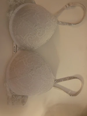 Vit push-up bh med spets och strass - Vit push-up bh med vadderade kupor och vacker spetsdetalj över hela bh:n. Axelbanden är dekorerade med strass för en extra glittrig touch. Bh:n har hakknäppning bak och mjuk insida för komfort. Säljer en likadan rosa bh. Priset kan diskuteras!