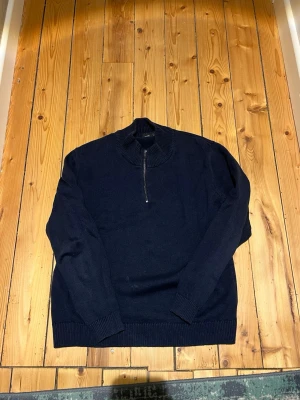 Mörkblå stickad halvzip tröja - Mörkblå stickad tröja med halv dragkedja och hög krage. Tröjan har långa ärmar och ribbade muddar vid ärmslut och nederkant. Perfekt för lager på lager och enkel att matcha med olika outfits.