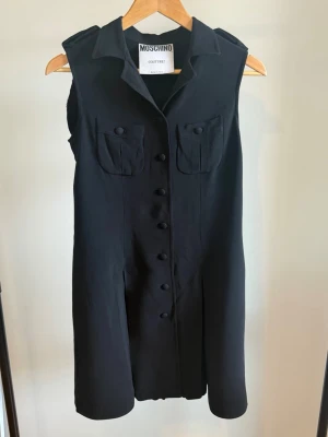Vintage klänning från Moschino Couture  - Ett så otroligt häftigt plagg från 90-talet. Det här är en äkta skräddad svart ärmlös klänning från Moschino Couture! (deras premiumlinje), tillverkad i Italien. Den är märkt USA 10 / UK 12 / IT 44, men eftersom det är vintage är den lite liten i storleken, så skicka ett meddelande om du vill ha exakta mått. Den har riktigt snygg plissering i ryggen och knäppta fickor framtill. Jättefint skick och superflatterande på.